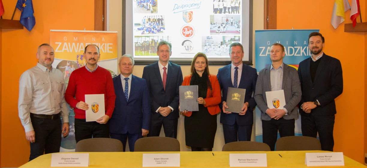 Grupa pracowników umysłowych w strojach formalnych, w tym garniturach, marynarkach i krawatach, stojąca i uśmiechająca się. Logo z tekstem "GMINA" widoczne.