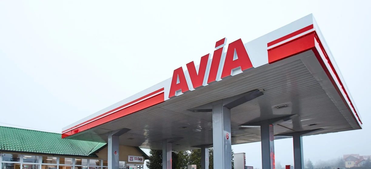 Samochód rodzinny zaparkowany na stacji benzynowej z widocznymi lusterkami samochodowymi i tabliczką rejestracyjną. Produkty marki AVÍA wyświetlane w tle.