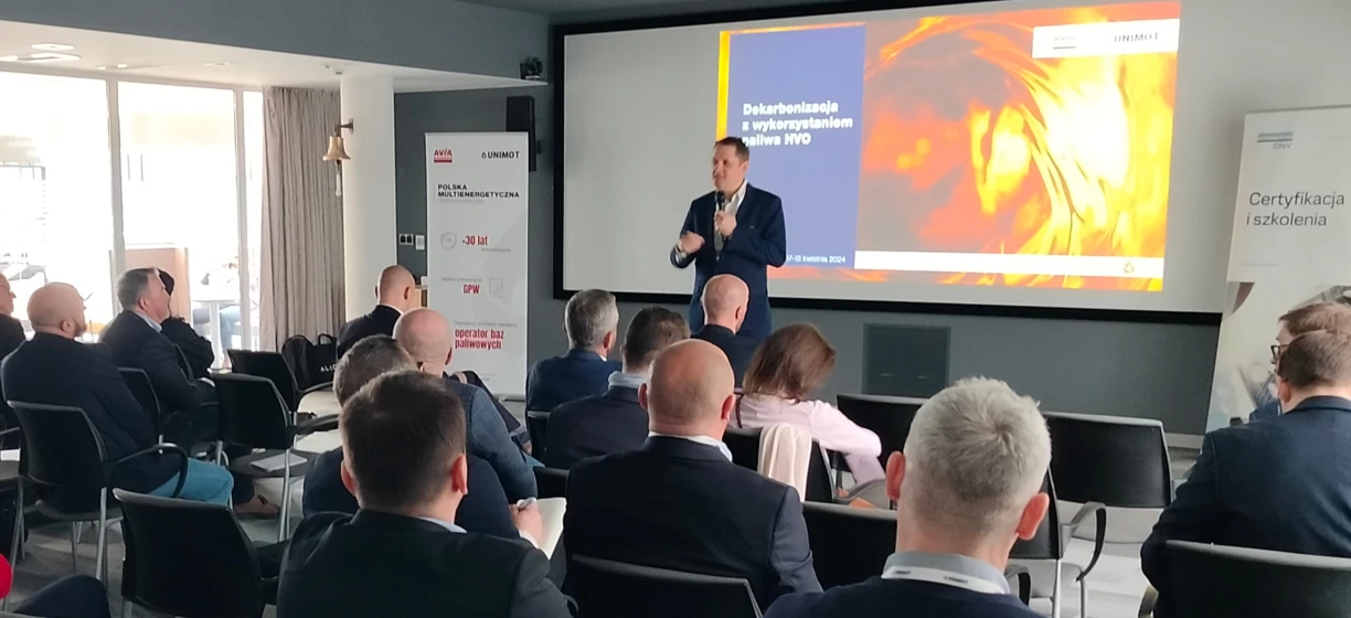 Tekst alternatywny obrazu: Osoba w garniturze prezentująca na urządzeniu wyświetlającym na wydarzeniu odzież wierzchnią. Interakcja i seminarium na temat najlepszych praktyk SEO w języku polskim.