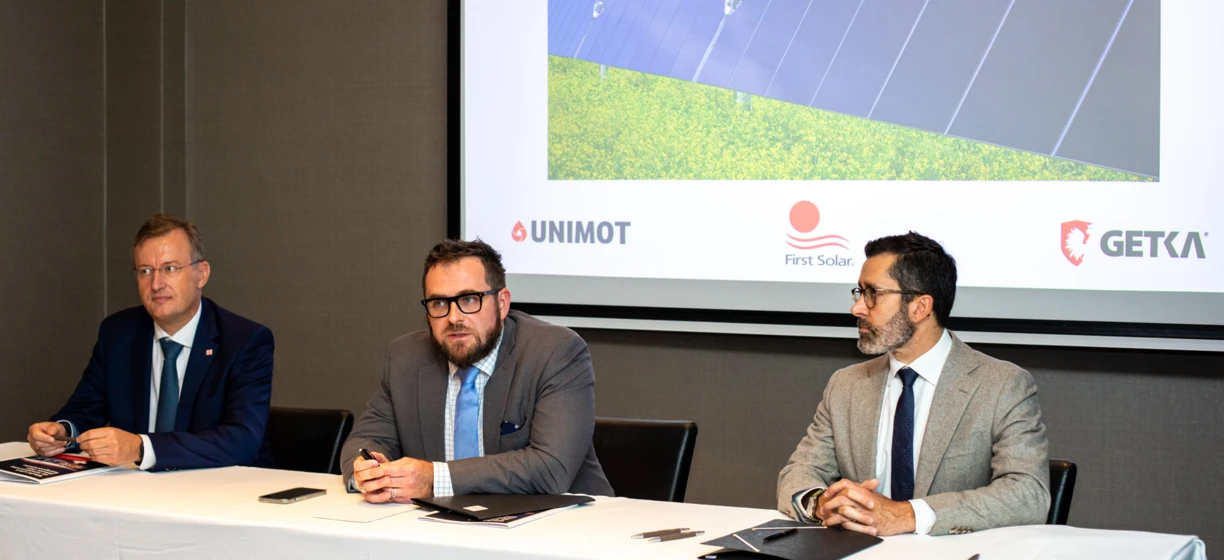 Pracownik biurowy w eleganckim ubraniu trzymający mikrofon na wydarzeniu z ubraniami, odzieżą zewnętrzną i urządzeniami elektronicznymi. Logo UNIMOT, GETKA i First Solar widoczne w widocznym miejscu.