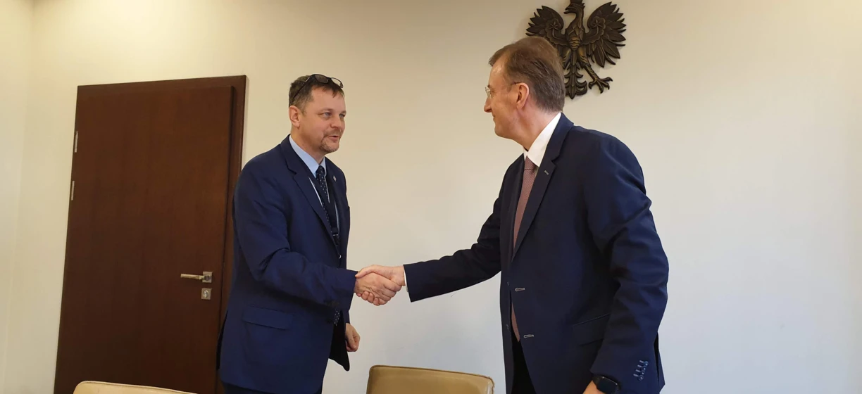 Mężczyzna w formalnym garniturze stoi przy wieszaku na płaszcze, poprawiając swoją krawat. Otaczają go meble, prezentując interakcję między modą a dekoracją.