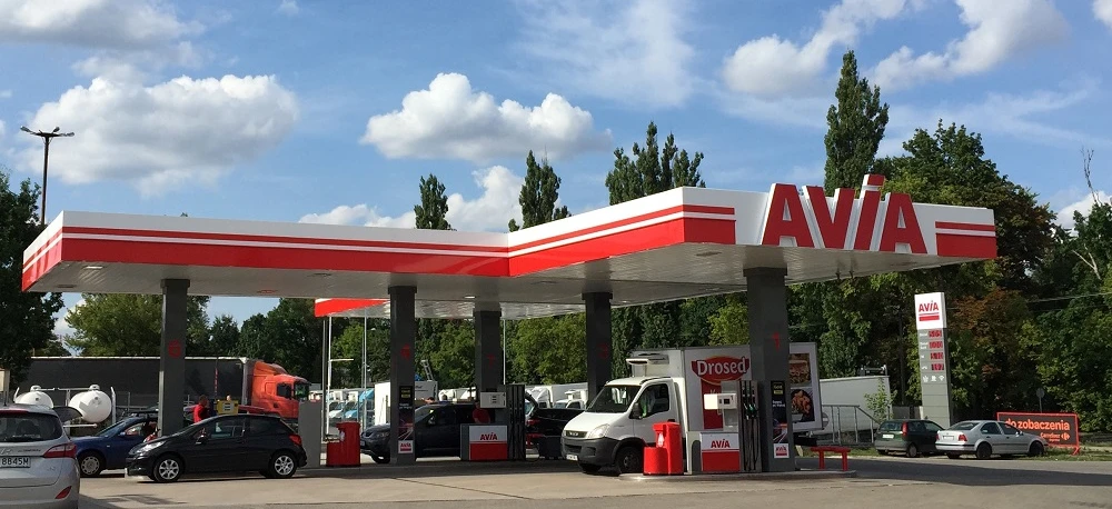 Stacja benzynowa z oznaczeniem AVIA, wyposażona w lampy parkingowe i lusterka boczne. Pojazd z blachą rejestracyjną Drosed tankuje paliwo przy dystrybutorze.