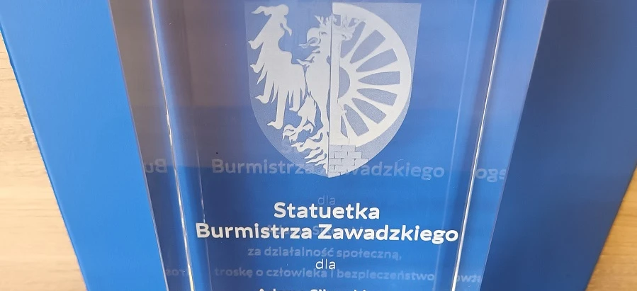 Obraz trofeum z napisem "Nagroda za działalność społeczną, troska o bezpieczeństwo ludzkie" wręczony przez burmistrza Zawadzkiego. Tło wykonane z transparentności i grafiki.