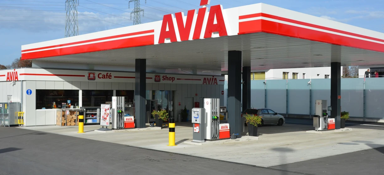 Stacja benzynowa z dystrybutorami paliwa, ciężarówkami i sklepem. Widoczne logo AVIA Café i SD Eibar. Ruchliwa scena motoryzacyjna z pompami benzynowymi.