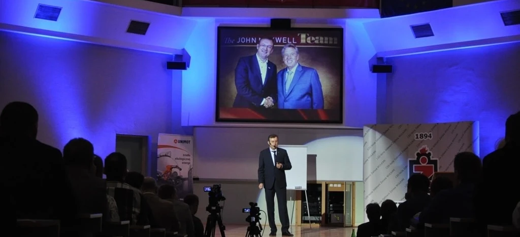 Rzecznik w garniturze wygłaszający przemówienie na wydarzeniu, stojący przy mównicy z urządzeniem do prezentacji i elektronicznymi urządzeniami w tle. Ubranie obejmuje płaszcz i odzież zewnętrzną. Prezentacja na konferencji QUNIMOT 2016.