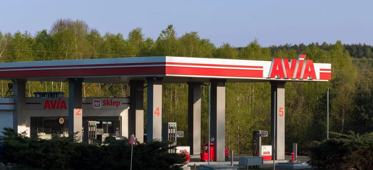 Obraz luksusowego samochodu zaparkowanego na stacji paliw z oznaczeniem AVIA. Samochód ma lśniący zderzak, kołpaki i lusterka boczne. Osobisty samochód luksusowy.