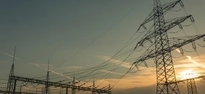 Linia przesyłowa i wieża przesyłowa na tle zachmurzonego nieba, symbolizująca elektryczność i użyteczność publiczną sieci elektroenergetycznej.