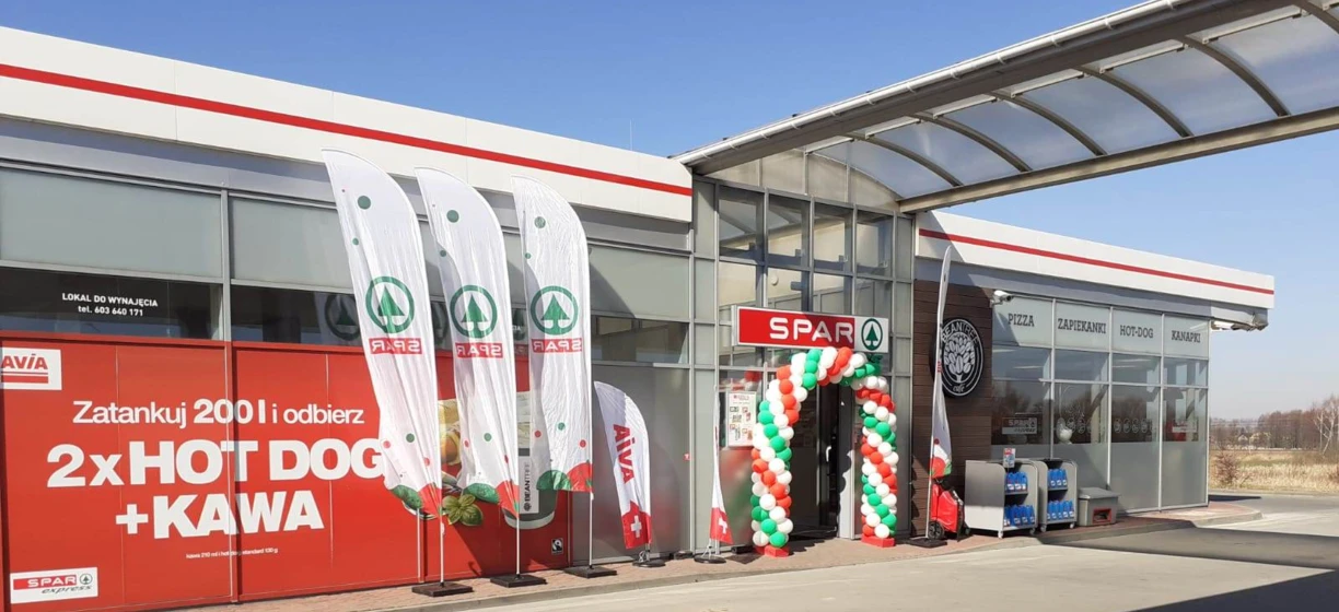 Logo AVIA na stacji benzynowej z reklamą hot dogów, kawy i pizzy. Lokal do wynajęcia z podanym numerem telefonu.