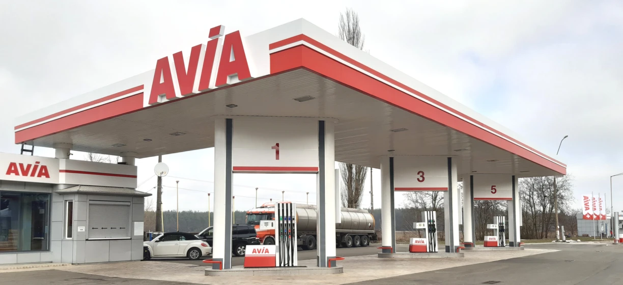 Samochód sportowo-użytkowy na stacji benzynowej z dyszą paliwową w zbiorniku paliwa samochodu, otoczony lusterkami samochodowymi i zderzakiem. Widoczna marka AVİA na osłonie piasty.