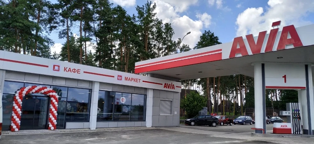 Obraz samochodu zaparkowanego na stacji paliw z otwartymi drzwiami. Osłona na piastę widoczna podczas tankowania. Znak po polsku dla "AVIA", "КАФЕ" i "MAPKET".