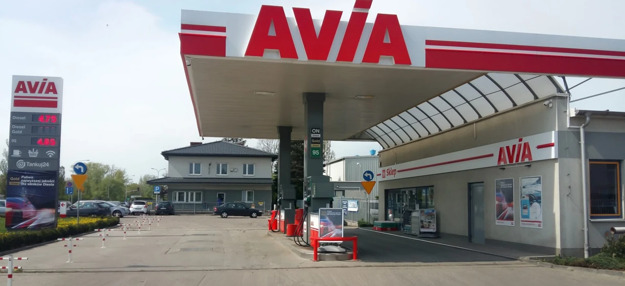 Obrazek opisowy: "Samochód z zapalonym przednim światłem postojowym i zderzakiem, z rejestracją i lusterkiem bocznym. Stacja paliwowa marki AVIA, sprzedająca olej napędowy o symbolu 95, dostępna w sklepie Tankuj24".