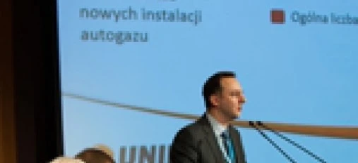 Rzecznik w formalnym stroju i płaszczu wygłasza przemówienie podczas wydarzenia, korzystając z sprzętu audio i mikrofonu. Tekst na obrazie podaje całkowitą liczbę konwersji na LPG w 2012 roku.