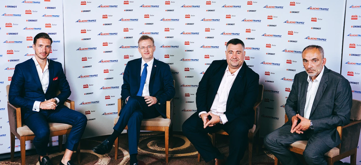 Mężczyzna w eleganckim ubraniu, stojący i uśmiechający się, ubrany w zestaw pracownika biurowego z marynarką, koszulą, krawatem i spodniami garniturowymi. Loga BLACHOTRAPEZ, AVIA Solar, UNIMOT na tle.