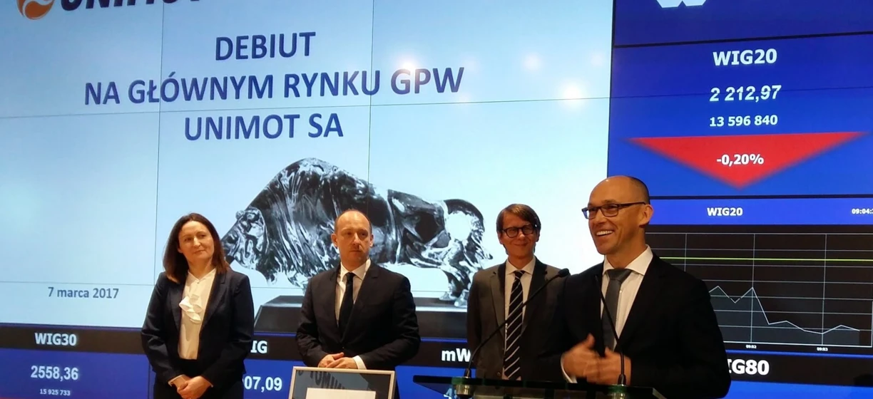 Tekst alternatywny: Profesjonalista w formalnym stroju białego kołnierza z płaszczem, marynarką i logo UNIMOT. Dane giełdowe w języku polskim na Giełdzie Papierów Wartościowych w Warszawie.