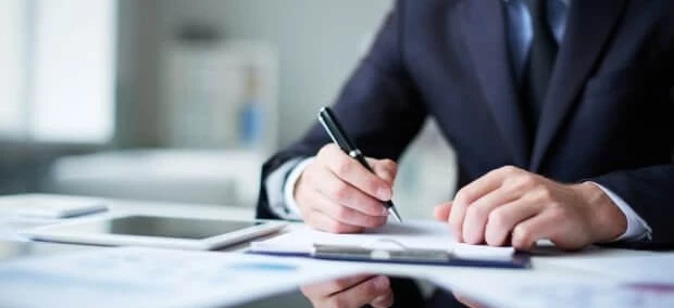 Pracownik umysłowy przy biurku z artykułami biurowymi, przyborami do pisania i papierem, skupiony na swojej pracy. Obraz reprezentuje profesjonalne środowisko i naukę.