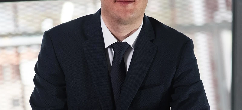 Osoba w garniturze i okularach pewnie stojąca z poważną miną. Modne okulary i stylowy strój wyrażają profesjonalizm.