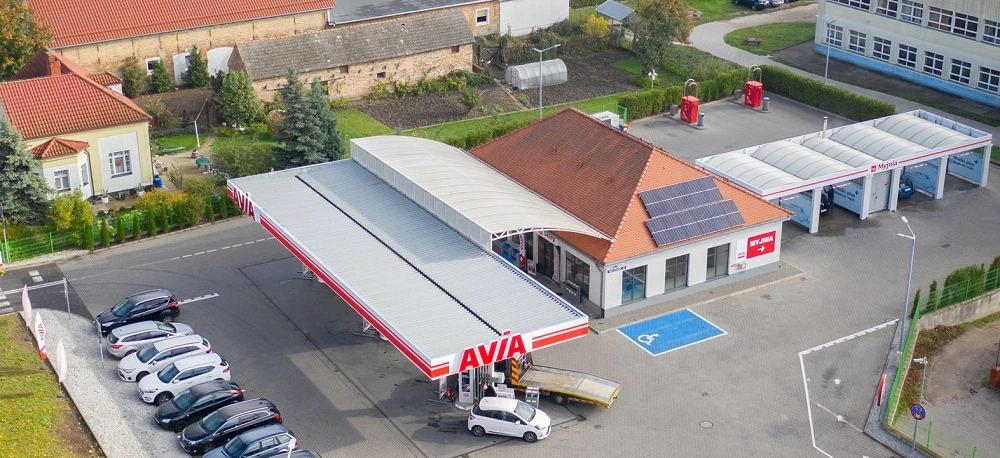 Ruchliwa stacja benzynowa z różnymi pojazdami zaparkowanymi, tankującymi paliwo pod jasnymi światłami parkingowymi i hamulcowymi. Zawiera samochody, ciężarówki i SUV-y z dystrybutorem w tle.