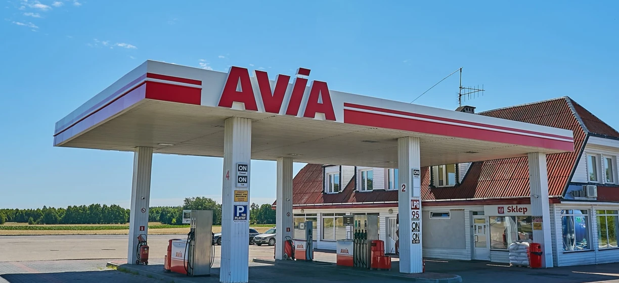 Odbicie stacji benzynowej w lusterku samochodowym z logo AVIA. Dystrybutor paliwa i benzyna dostępna. Lusterko wsteczne pokazuje logo AVIA.