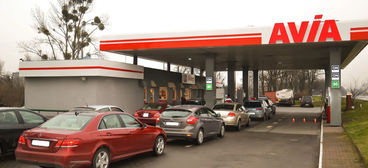 Obraz samochodu rodinnego zaparkowanego na stacji benzynowej, z błyszczącym lustrem samochodu i tablicą rejestracyjną. Drzwi i zderzak samochodu są widoczne, wraz z lusterkiem bocznym samochodu.