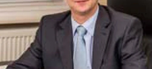 Osoba biznesowa w eleganckim garniturze i krawacie uśmiechająca się z profesjonalną mimiką twarzy. Pracownik biurowy w koszuli i marynarce.