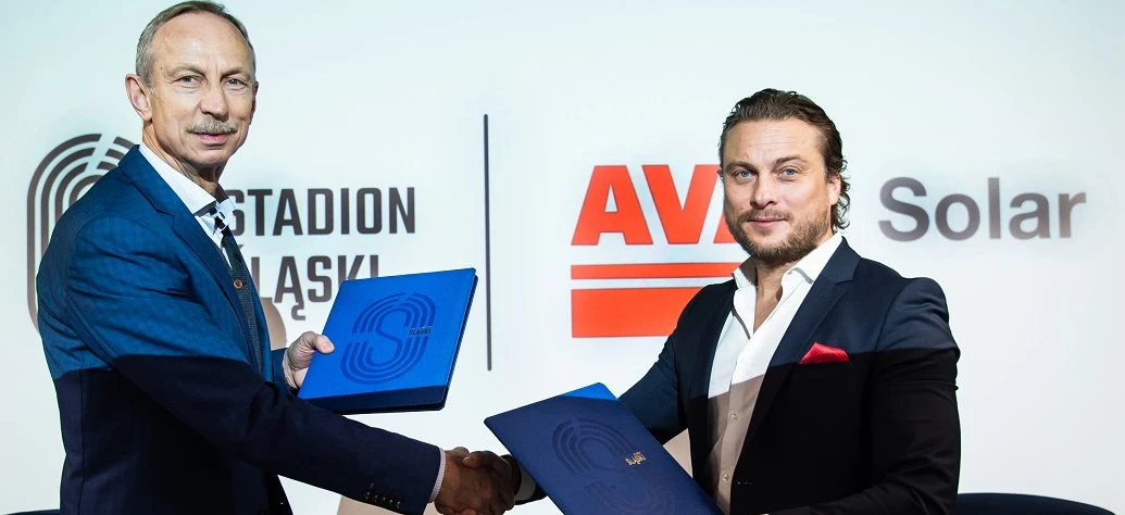 Pracownik umysłowy w eleganckim garniturze i marynarce uśmiecha się obok logo reklamowego AV Solar na STADIONIE. Promując profesjonalny styl i czyste rozwiązania energetyczne.