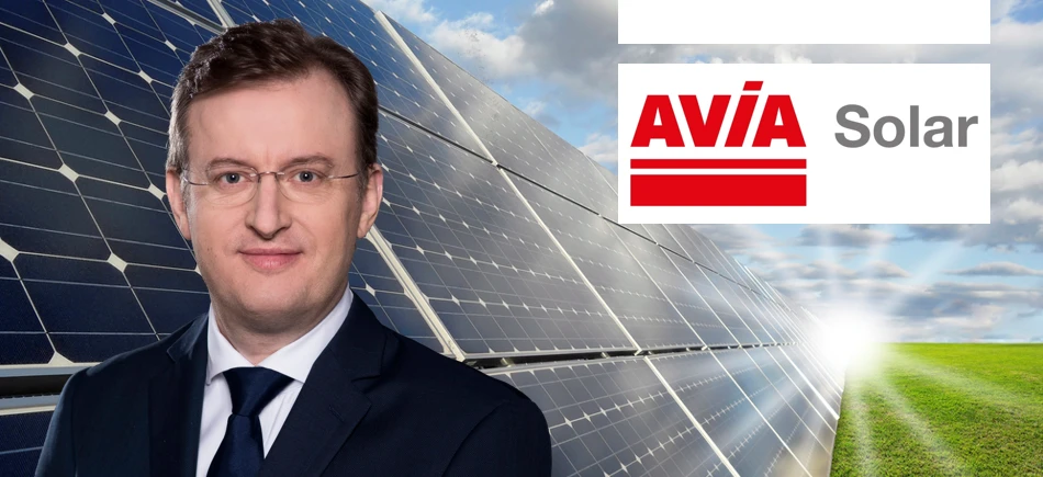 Pracownik umysłowy w garniturze i krawacie stoi obok panelu słonecznego z reklamą UNIMOT, firmy specjalizującej się w energii słonecznej. W tle widoczne jest również AVİA Solar.