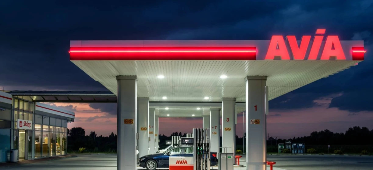 Obraz nowoczesnej stacji benzynowej z dystrybutorem paliwa i sklepem sprzedającym produkty, takie jak benzyna. Widoczne są wyraźnie oznaczenia marki AVIA.