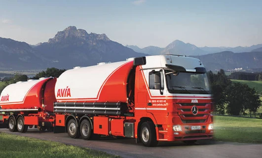 Pojazd dostawczy z logo AVÍA zaparkowany przed zbiornikiem magazynowym. Ciężarówka naczepowa z oznaczeniem AVIA oraz cechami Mercedes-Benz. Idealna do transportu towarowego.
