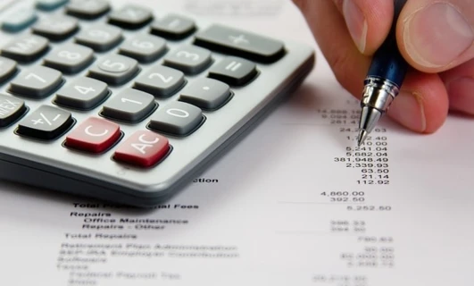 Biurko biurowe z kalkulatorem, papierem i innymi artykułami biurowymi do obliczania całkowitych kosztów i wydatków. Słowa kluczowe obejmują sprzęt biurowy, artykuły papiernicze, dokumenty i przybory do pisania. Podsumowanie liczb na potrzeby utrzymania biura oraz napraw.