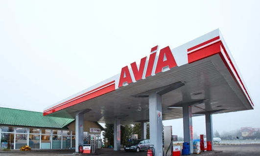Samochód rodzinny zaparkowany na stacji benzynowej z widocznymi lusterkami samochodowymi i tabliczką rejestracyjną. Produkty marki AVÍA wyświetlane w tle.