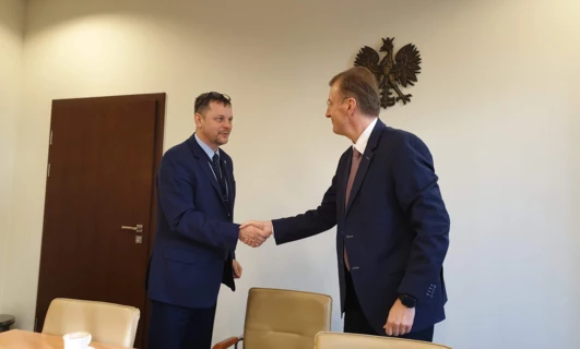 Mężczyzna w formalnym garniturze stoi przy wieszaku na płaszcze, poprawiając swoją krawat. Otaczają go meble, prezentując interakcję między modą a dekoracją.