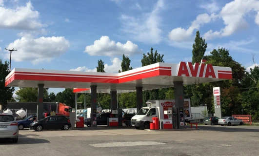 Stacja benzynowa z oznaczeniem AVIA, wyposażona w lampy parkingowe i lusterka boczne. Pojazd z blachą rejestracyjną Drosed tankuje paliwo przy dystrybutorze.