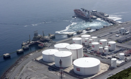 Rozgardiaszony krajobraz przemysłowy z zbiornikami magazynowymi, transportem towarowym i elektrownią przy porcie. Loga AVIA i SD Eibar są wyraźnie widoczne.