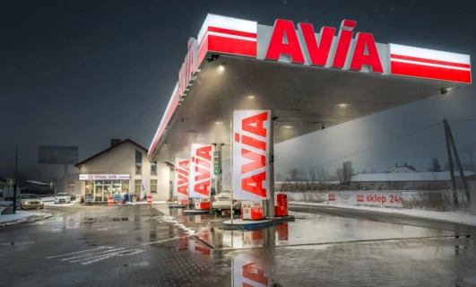 Cysternowa ciężarówka paliwowa zaparkowana na stacji benzynowej czynnej 24 godziny na dobę (Sklep 24h) z widocznym logo AVÍA na zderzaku.