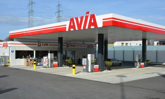 Stacja benzynowa z dystrybutorami paliwa, ciężarówkami i sklepem. Widoczne logo AVIA Café i SD Eibar. Ruchliwa scena motoryzacyjna z pompami benzynowymi.