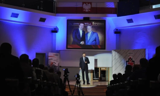 Rzecznik w garniturze wygłaszający przemówienie na wydarzeniu, stojący przy mównicy z urządzeniem do prezentacji i elektronicznymi urządzeniami w tle. Ubranie obejmuje płaszcz i odzież zewnętrzną. Prezentacja na konferencji QUNIMOT 2016.