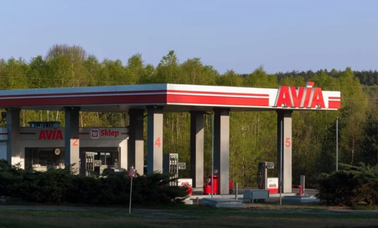 Obraz luksusowego samochodu zaparkowanego na stacji paliw z oznaczeniem AVIA. Samochód ma lśniący zderzak, kołpaki i lusterka boczne. Osobisty samochód luksusowy.