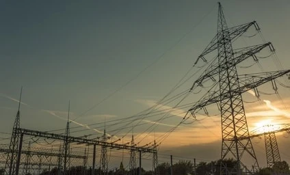 Linia przesyłowa i wieża przesyłowa na tle zachmurzonego nieba, symbolizująca elektryczność i użyteczność publiczną sieci elektroenergetycznej.