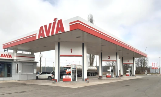 Samochód sportowo-użytkowy na stacji benzynowej z dyszą paliwową w zbiorniku paliwa samochodu, otoczony lusterkami samochodowymi i zderzakiem. Widoczna marka AVİA na osłonie piasty.