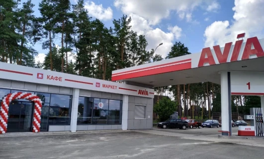 Obraz samochodu zaparkowanego na stacji paliw z otwartymi drzwiami. Osłona na piastę widoczna podczas tankowania. Znak po polsku dla "AVIA", "КАФЕ" i "MAPKET".