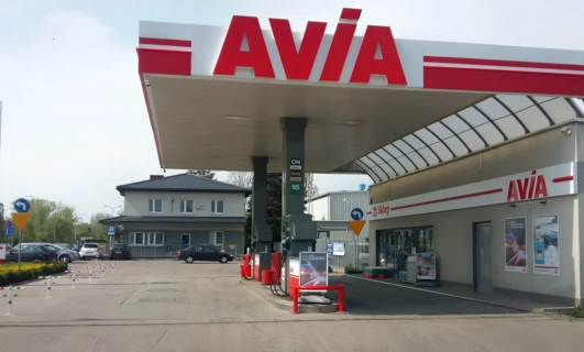 Obrazek opisowy: "Samochód z zapalonym przednim światłem postojowym i zderzakiem, z rejestracją i lusterkiem bocznym. Stacja paliwowa marki AVIA, sprzedająca olej napędowy o symbolu 95, dostępna w sklepie Tankuj24".