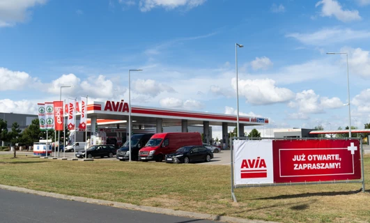 Lusterko boczne ciężarówki z reklamą AVIA, SPAR i SD Eibar na stacji benzynowej. Znak "AVIA Truck" z napisem po polsku "JUŻ OTWARTE, ZAPRASZAMY!" w tle.