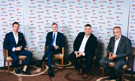 Mężczyzna w eleganckim ubraniu, stojący i uśmiechający się, ubrany w zestaw pracownika biurowego z marynarką, koszulą, krawatem i spodniami garniturowymi. Loga BLACHOTRAPEZ, AVIA Solar, UNIMOT na tle.