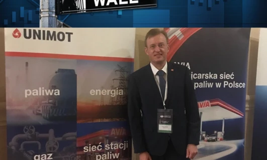 Pracownik umysłowy w garniturze i marynarce stoi obok banerów reklamowych. Słowa kluczowe: Praca, biznesmen, logo, UNIMOT, Wall St.