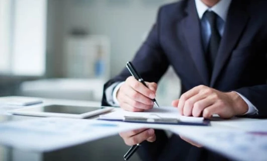 Pracownik umysłowy przy biurku z artykułami biurowymi, przyborami do pisania i papierem, skupiony na swojej pracy. Obraz reprezentuje profesjonalne środowisko i naukę.