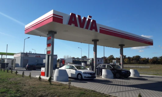 Widok z tyłu samochodu subkompaktowego na stacji benzynowej, z odbiciem lusterka bocznego i zderzaka widocznym. Pokazane są różne ceny paliwa i marka AVIA.