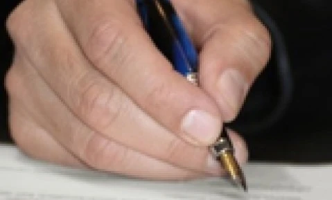 Ręka osoby trzymająca długopis nad kawałkiem papieru na biurku z przyborami biurowymi. Pisanie z nadgarstkiem opartym na stole.