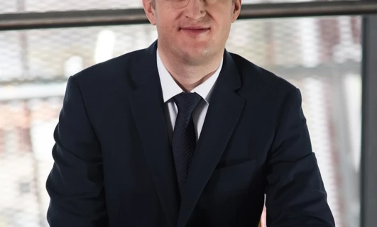 Osoba w garniturze i okularach pewnie stojąca z poważną miną. Modne okulary i stylowy strój wyrażają profesjonalizm.