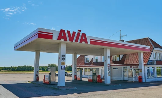 Odbicie stacji benzynowej w lusterku samochodowym z logo AVIA. Dystrybutor paliwa i benzyna dostępna. Lusterko wsteczne pokazuje logo AVIA.
