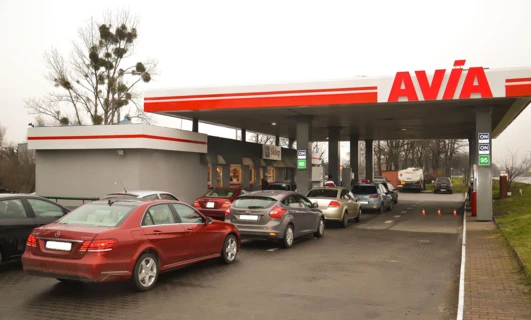 Obraz samochodu rodinnego zaparkowanego na stacji benzynowej, z błyszczącym lustrem samochodu i tablicą rejestracyjną. Drzwi i zderzak samochodu są widoczne, wraz z lusterkiem bocznym samochodu.